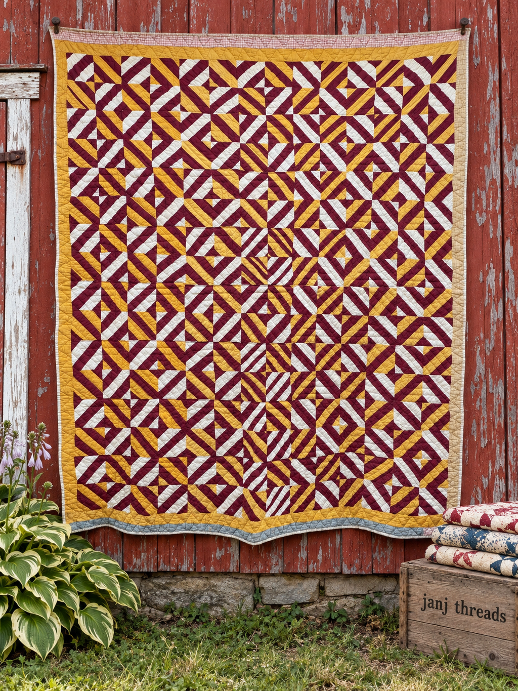 A large quilted textile with a repeating geometric pattern in burgundy, mustard yellow, and off-white hangs against a weathered red wooden barn wall. A small plant is at the lower left, and a wooden crate with folded quilts sits at the lower right.