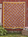A large quilted textile with a repeating geometric pattern in burgundy, mustard yellow, and off-white hangs against a weathered red wooden barn wall. A small plant is at the lower left, and a wooden crate with folded quilts sits at the lower right.