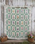 A vintage-style floral patchwork quilt hanging on a weathered white wooden wall, with a basket of threads on the left and a flower basket on the right.