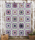 A vintag Blazing Star  handmade quilt featuring a grid of starburst patterns in lavender, mint green, pink, navy, and beige, hanging on a rustic white-painted wooden wall. Two wicker baskets with floral arrangements sit on the ground at the bottom corners, and the brand name 'Janji Threads' is printed on the left basket.