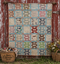 Broken Path Variation quilt, a vintage-style patchwork quilt with a grid of geometric and floral blocks in muted pastel and earthy tones, hanging on a rustic red wooden barn door. A wicker basket of spools of thread sits on the left, and a basket of wildflowers sits on the right, with grass visible at the bottom.