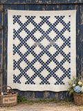 A large quilted patchwork blanket with a navy blue and cream geometric lattice pattern is hung against a weathered dark blue wooden barn wall. A woven basket of thread spools sits on the left, and a small flower pot with daisies and blue hydrangeas sits on the right.
