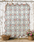 A handmade quilted patchwork quilt with a scalloped edge, featuring repeating interlocking circle patterns in pastel colors, hanging against a weathered white wooden wall. A wicker basket labeled 'Jan Threads' with eggs sits on the left, and a wicker basket with pink flowers sits on the right.