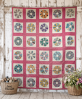 A handmade Dresden Plate quilt featuring a grid of colorful circular patchwork medallions on a pink background, hung against a rustic white wooden wall with baskets of flowers and yarn at the bottom.