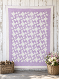 A lavender and white drunkard's path quilted wall hanging or tapestry with a repeating geometric pinwheel pattern, displayed against a rustic white wood wall with a basket of wood shavings and a flower basket on either side.