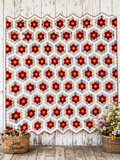 A large quilted textile with an allover floral patchwork pattern hangs against a rustic white wood wall. The design features red and dark red flowers with yellow centers, surrounded by blue, green, and orange floral motifs on a white background. Grandmother's Flower Garden Quilt. 