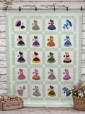 A handmade quilt hanging on a rustic white wooden wall, featuring a grid of embroidered dresses in various colors on a pale green and white checkered background. A wooden crate labeled 'Janj Threads' and a basket of flowers sit at the bottom.