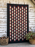A large patchwork quilt with vertical columns of cream and red fabric blocks on a black background, displayed against a rustic white wood wall. A wicker basket labeled 'Janj Threads' sits at the bottom left, and a flower basket is at the bottom right.