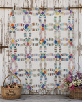 A colorful quilted patchwork blanket with interlocking circular patterns hangs on a rustic white wooden wall. The quilt features a white base with multicolored geometric and striped segments in shades of blue, pink, yellow, green, and orange.